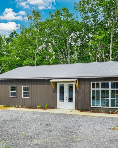 Modern metal-sided cabin with metal roof, yellow trim, and white doors surrounded by tall trees and gravel driveway.
