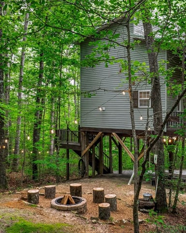 Elevated treehouse cabin with wraparound deck surrounded by dense forest, fire pit and log seating below, hammock strung between trees.