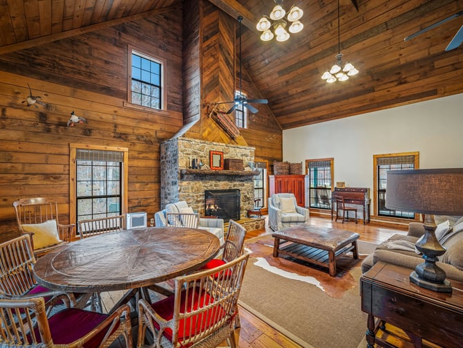 Spacious log cabin interior with stone fireplace, dining table, seating areas, and vaulted wood ceiling with pendant lights