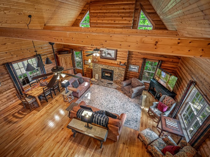 Overhead view of log cabin interior with wood beams, fireplace, dining area, and multiple seating zones with large windows