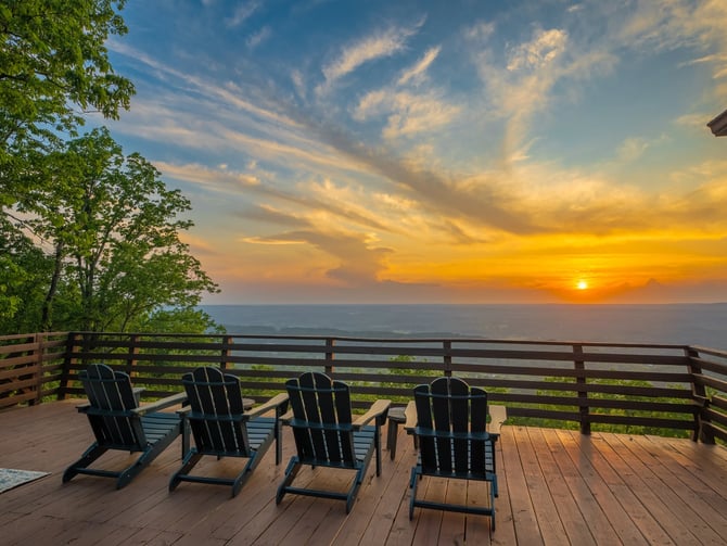 Wooden deck with four black Adirondack chairs overlooking ocean at sunset, wooden railing and green trees